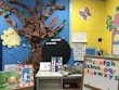 A colorful classroom corner decorated with educational materials and artwork. A tree made of brown paper with a bright yellow paper sun on a blue background. Alphabet letters displayed on a board with multicolored magnets. Several educational posters feature drawings and letters, and a plush toy lies on a white shelf. Various cut-out images and decorations create a child-friendly and engaging learning environment.
