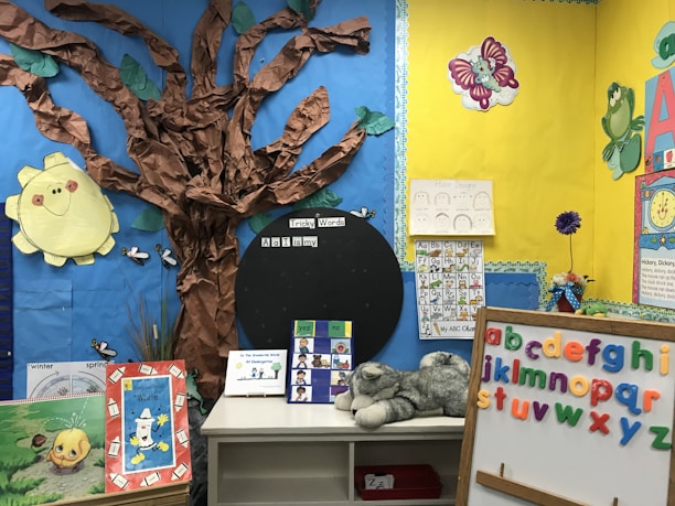 A colorful classroom corner decorated with educational materials and artwork. A tree made of brown paper with a bright yellow paper sun on a blue background. Alphabet letters displayed on a board with multicolored magnets. Several educational posters feature drawings and letters, and a plush toy lies on a white shelf. Various cut-out images and decorations create a child-friendly and engaging learning environment.