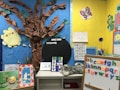 A colorful classroom corner decorated with educational materials and artwork. A tree made of brown paper with a bright yellow paper sun on a blue background. Alphabet letters displayed on a board with multicolored magnets. Several educational posters feature drawings and letters, and a plush toy lies on a white shelf. Various cut-out images and decorations create a child-friendly and engaging learning environment.