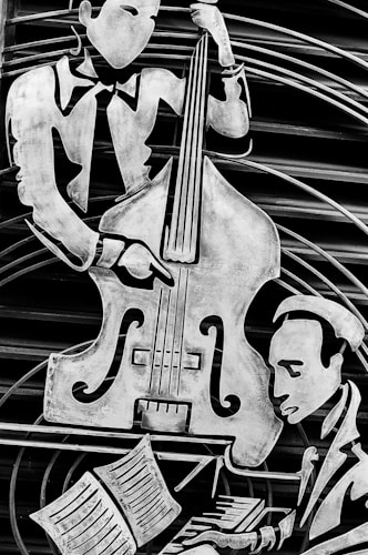 A black and white relief sculpture featuring two musicians. One is playing a large string instrument resembling a double bass, wearing formal attire with a tie. The other musician is playing a piano, sitting in front of an open musical score. The artwork uses stylized, angular forms and lines to convey a sense of movement.