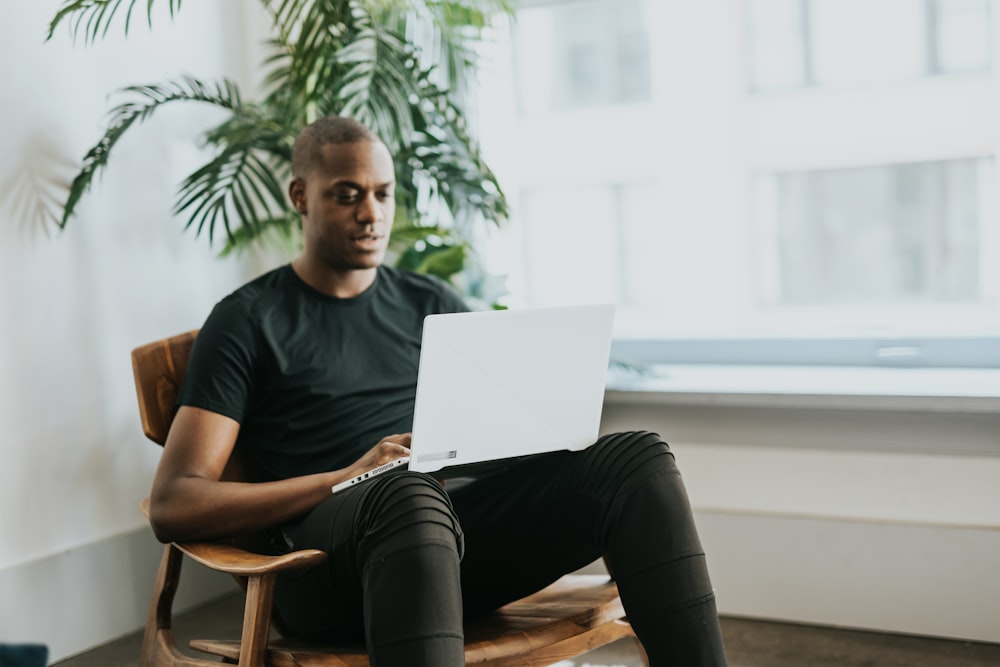 검은 크루 넥 티셔츠를 입은 남자는 노트북을 사용하여 갈색 나무 의자에 앉아