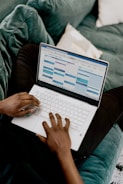 A person is sitting on a green couch, using a white laptop with a calendar application open on the screen.