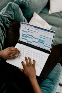 A person is sitting on a green couch, using a white laptop with a calendar application open on the screen.