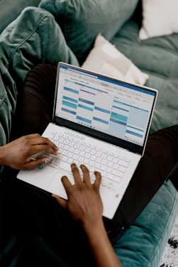 A person is sitting on a green couch, using a white laptop with a calendar application open on the screen.