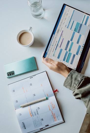 A workspace setup with a tablet displaying a calendar, a cup of coffee, a smartphone, a pen, and an open planner on a desk. The calendar on the tablet shows a digital scheduling app, while the planner has handwritten notes.