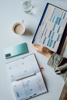A workspace setup with a tablet displaying a calendar, a cup of coffee, a smartphone, a pen, and an open planner on a desk. The calendar on the tablet shows a digital scheduling app, while the planner has handwritten notes.