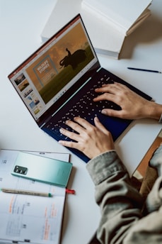 A person is typing on a laptop with a presentation slide showing a wildlife theme on the screen. Nearby, a smartphone and a planner with notes are placed on the desk. The setting suggests a work or study environment, with a pen placed beside the laptop.