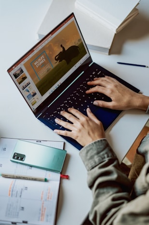 A person is typing on a laptop with a presentation slide showing a wildlife theme on the screen. Nearby, a smartphone and a planner with notes are placed on the desk. The setting suggests a work or study environment, with a pen placed beside the laptop.