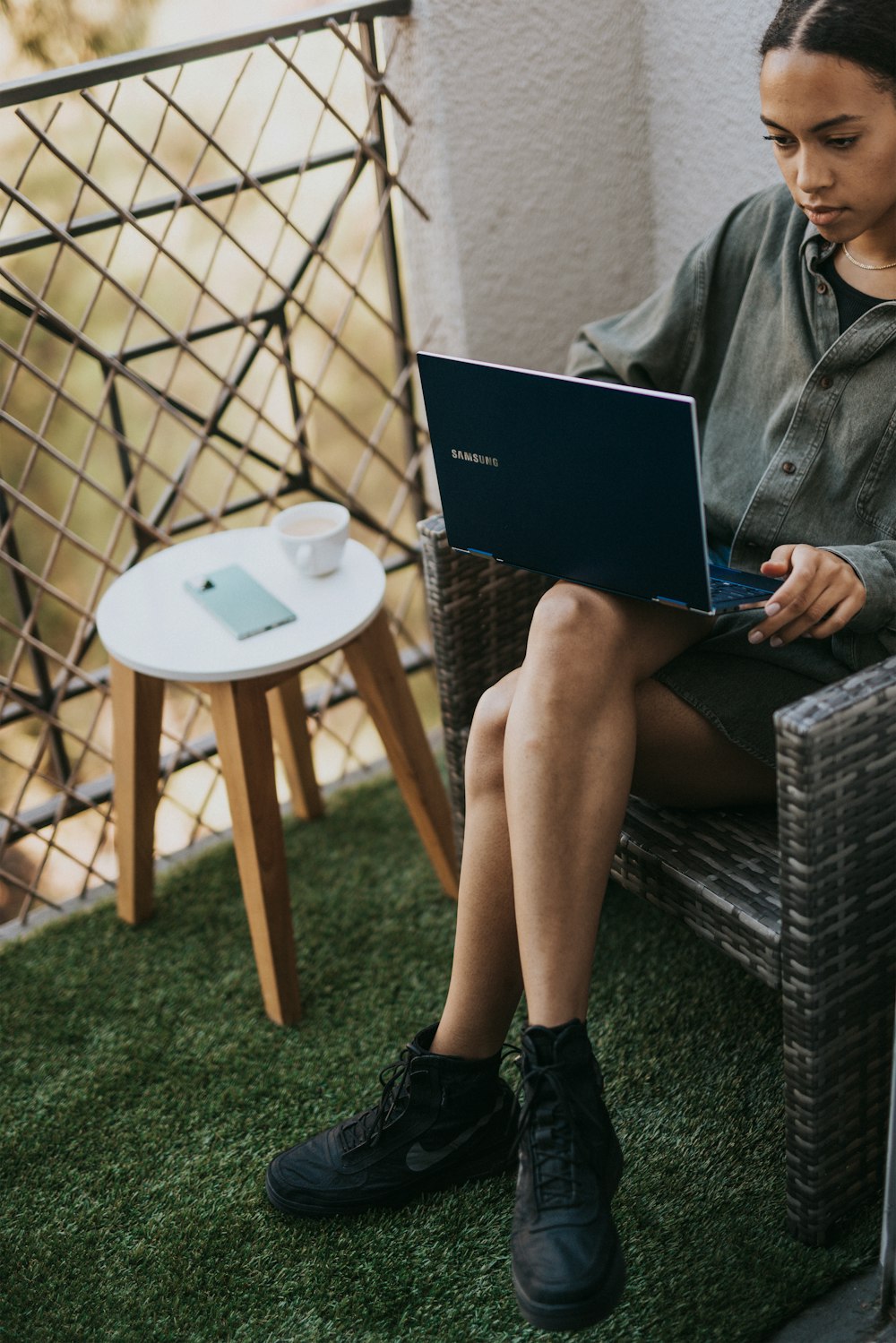 woman in gray coat sitting on black wicker chair using black laptop computer