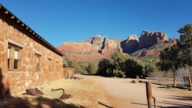 A rustic stone building is situated in a sunlit landscape with towering red rock formations in the background. The area is surrounded by desert vegetation and a clear blue sky overhead.