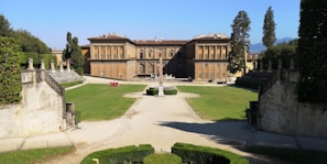 A large historic building with a symmetrical facade, featuring multiple windows and a central entrance. The building is surrounded by a well-maintained lawn with a gravel path leading to an obelisk centerpiece. The garden is bordered by stone walls and tall cypress trees.