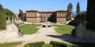 A large historic building with a symmetrical facade, featuring multiple windows and a central entrance. The building is surrounded by a well-maintained lawn with a gravel path leading to an obelisk centerpiece. The garden is bordered by stone walls and tall cypress trees.