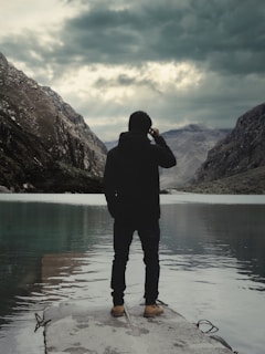 A person stands on a concrete platform at the edge of a serene lake, surrounded by mountains under a dramatic, cloudy sky.
