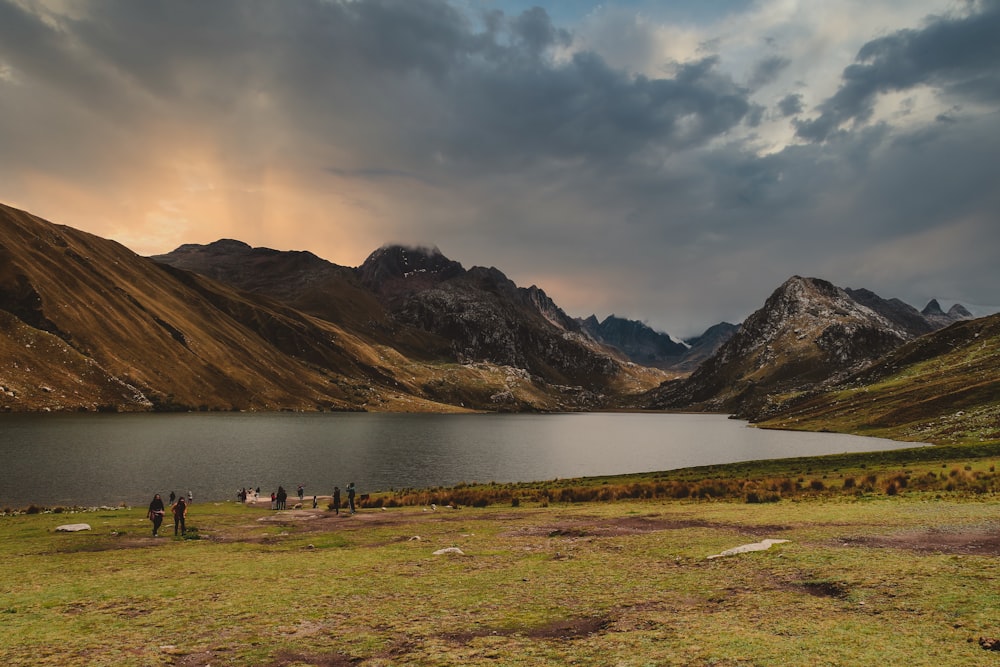 people standing on green grass field near lake and mountains under white clouds and blue sky