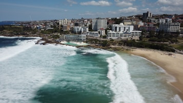 A coastal view featuring a sandy beach and an adjacent swimming pool bordered by a rocky coastline. Tall buildings and residential complexes line the shore in the background. The ocean waves break energetically onto the shore, while the sky is dotted with scattered clouds.