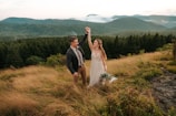 A couple stands on a grassy hilltop surrounded by mountains and lush forests. The woman is wearing a white dress and holding a bouquet of flowers, while the man wears a suit and raises her hand gently. The background features rolling hills covered in green foliage under a cloudy sky, creating a serene and romantic atmosphere.