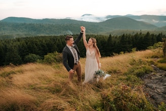 A couple stands on a grassy hilltop surrounded by mountains and lush forests. The woman is wearing a white dress and holding a bouquet of flowers, while the man wears a suit and raises her hand gently. The background features rolling hills covered in green foliage under a cloudy sky, creating a serene and romantic atmosphere.