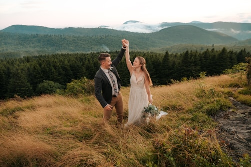 A couple stands on a grassy hilltop surrounded by mountains and lush forests. The woman is wearing a white dress and holding a bouquet of flowers, while the man wears a suit and raises her hand gently. The background features rolling hills covered in green foliage under a cloudy sky, creating a serene and romantic atmosphere.