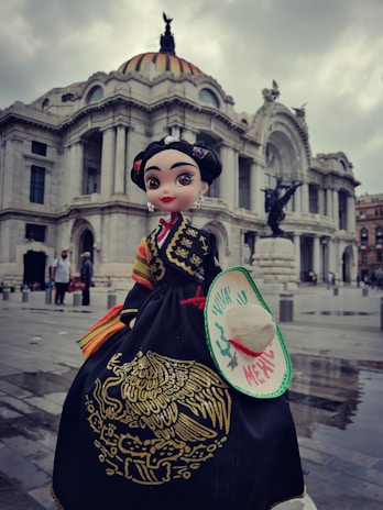 A doll dressed in traditional Mexican attire stands in front of a grand, historic building with distinctive architectural features. The doll wears a black dress with gold embroidery and colorful accents, and holds a small hat with the words 'Viva la Mexico' on it. The background features a cloudy sky and a few people walking in the vicinity.