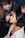 A makeup artist is applying makeup to a woman's face. The artist is wearing glasses and a black leather jacket, while the woman being styled has short black hair and is wearing a light-colored jacket. The scene appears focused and intimate, capturing the artist's precision and the woman's trust.