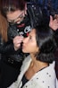 A makeup artist is applying makeup to a woman's face. The artist is wearing glasses and a black leather jacket, while the woman being styled has short black hair and is wearing a light-colored jacket. The scene appears focused and intimate, capturing the artist's precision and the woman's trust.