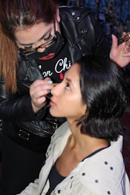 A makeup artist is applying makeup to a woman's face. The artist is wearing glasses and a black leather jacket, while the woman being styled has short black hair and is wearing a light-colored jacket. The scene appears focused and intimate, capturing the artist's precision and the woman's trust.