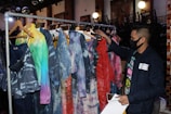 A person wearing a black face mask browses through a rack of clothing featuring tie-dye and colorful patterns. The garments, hanging on wooden hangers, display various hues, including blue, red, and green shades. The setting appears to be indoors, possibly a fashion store or exhibition space, with chairs stacked in the background and soft lighting contributing to a cozy atmosphere.
