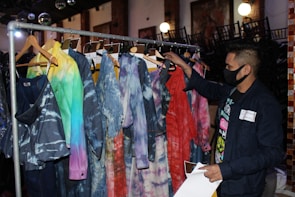 A person wearing a black face mask browses through a rack of clothing featuring tie-dye and colorful patterns. The garments, hanging on wooden hangers, display various hues, including blue, red, and green shades. The setting appears to be indoors, possibly a fashion store or exhibition space, with chairs stacked in the background and soft lighting contributing to a cozy atmosphere.