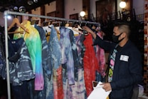 A person wearing a black face mask browses through a rack of clothing featuring tie-dye and colorful patterns. The garments, hanging on wooden hangers, display various hues, including blue, red, and green shades. The setting appears to be indoors, possibly a fashion store or exhibition space, with chairs stacked in the background and soft lighting contributing to a cozy atmosphere.