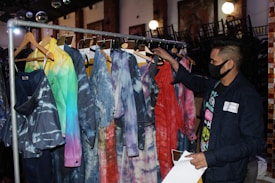 A person wearing a black face mask browses through a rack of clothing featuring tie-dye and colorful patterns. The garments, hanging on wooden hangers, display various hues, including blue, red, and green shades. The setting appears to be indoors, possibly a fashion store or exhibition space, with chairs stacked in the background and soft lighting contributing to a cozy atmosphere.