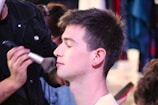 A person is having makeup applied by a makeup artist, with their eyes closed and a brush near their cheek. The focus is on the serene expression of the person receiving the makeup, while the artist is partly in view, concentrating on the task.