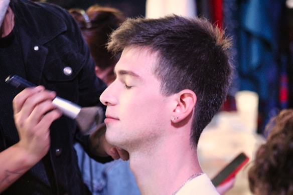 A person is having makeup applied by a makeup artist, with their eyes closed and a brush near their cheek. The focus is on the serene expression of the person receiving the makeup, while the artist is partly in view, concentrating on the task.