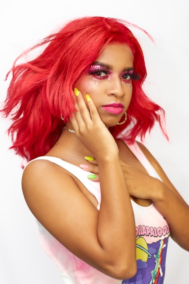 A person with bright red hair styled in waves, wearing bold pink makeup with sparkly accents on the eyelids. They have long yellow nails and are dressed in a sleeveless top featuring a colorful design. The background is plain white, highlighting the vivid colors of the hair and makeup.