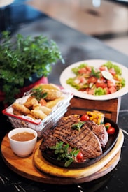 An appetizing meal is presented, consisting of a grilled steak garnished with fresh herbs and cherry tomatoes on a hot plate. To accompany the steak, there is a small cup of sauce on a round wooden serving board. French fries are served in a metal basket with a red and white patterned cloth. In the background, there is a fresh salad with lettuce, tomatoes, and lime slices on a white plate. A lush green plant adds a touch of freshness to the setting.
