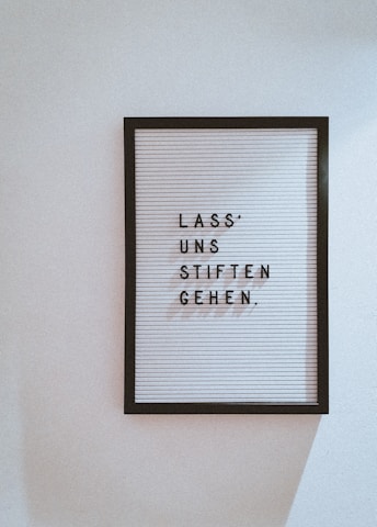 A black framed letter board is mounted on a white wall. The board features horizontal grooves with movable black letters spelling 'LASS' UNS STIFTEN GEHEN.'