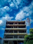 A multi-story concrete building under construction with exposed floors and columns. Several workers can be seen on the upper levels, engaged in construction activities. The sky is partly cloudy, and a telecommunications tower is visible to the side of the building.