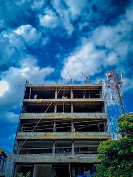 A multi-story concrete building under construction with exposed floors and columns. Several workers can be seen on the upper levels, engaged in construction activities. The sky is partly cloudy, and a telecommunications tower is visible to the side of the building.