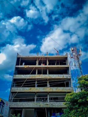 A multi-story concrete building under construction with exposed floors and columns. Several workers can be seen on the upper levels, engaged in construction activities. The sky is partly cloudy, and a telecommunications tower is visible to the side of the building.