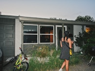 A girl with braided hair and wearing a black polka dot dress stands in front of a house. The house has large windows and a front door with frosted glass. There is a bicycle parked next to a closed garage door, and ornamental grass grows in front of the windows. It's early evening, and the interior lights are on, casting a warm glow through the windows.