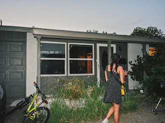 A girl with braided hair and wearing a black polka dot dress stands in front of a house. The house has large windows and a front door with frosted glass. There is a bicycle parked next to a closed garage door, and ornamental grass grows in front of the windows. It's early evening, and the interior lights are on, casting a warm glow through the windows.