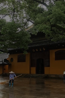 A person wearing a wide-brimmed hat and rubber boots is sweeping a wet stone courtyard in front of a traditional building with orange walls and wooden details. Overhead, large green trees create a canopy, adding a serene atmosphere to the scene.