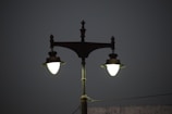 A vintage-style double street lamp is lit against a dark, plain background. The lamp post is ornate, with decorative elements at the top and a greenish hue to the metal. The lights are glowing brightly, contrasting with the dim surroundings.