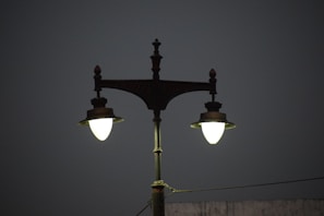 A vintage-style double street lamp is lit against a dark, plain background. The lamp post is ornate, with decorative elements at the top and a greenish hue to the metal. The lights are glowing brightly, contrasting with the dim surroundings.