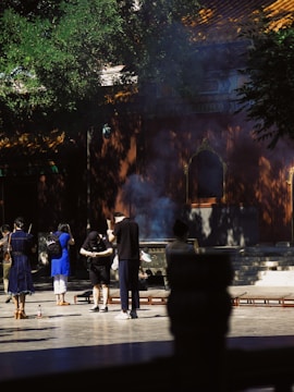A group of people are engaged in a traditional or ritualistic activity at a historical or cultural site. Shadows of trees and building details create an interplay of light and shadow. Some individuals appear to be holding incense sticks as smoke wafts into the air. The setting includes a stone floor and a structure with ornate architectural details in the background.