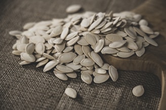 A pile of white, oval-shaped seeds is spread out on a rustic wooden surface, with a textured brown fabric underneath. The seeds appear to be pumpkin seeds, creating a natural and earthy ambiance.