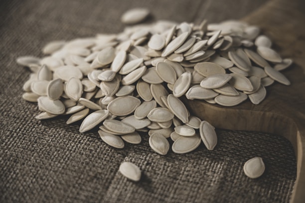 A pile of white, oval-shaped seeds is spread out on a rustic wooden surface, with a textured brown fabric underneath. The seeds appear to be pumpkin seeds, creating a natural and earthy ambiance.