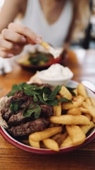 A plate of grilled meat accompanied by French fries, garnished with fresh mint. There is a bowl of white dipping sauce next to it, and a hand reaching to dip a fry into the sauce. The setting suggests a casual dining environment.