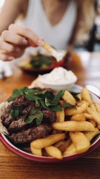 A plate of grilled meat accompanied by French fries, garnished with fresh mint. There is a bowl of white dipping sauce next to it, and a hand reaching to dip a fry into the sauce. The setting suggests a casual dining environment.