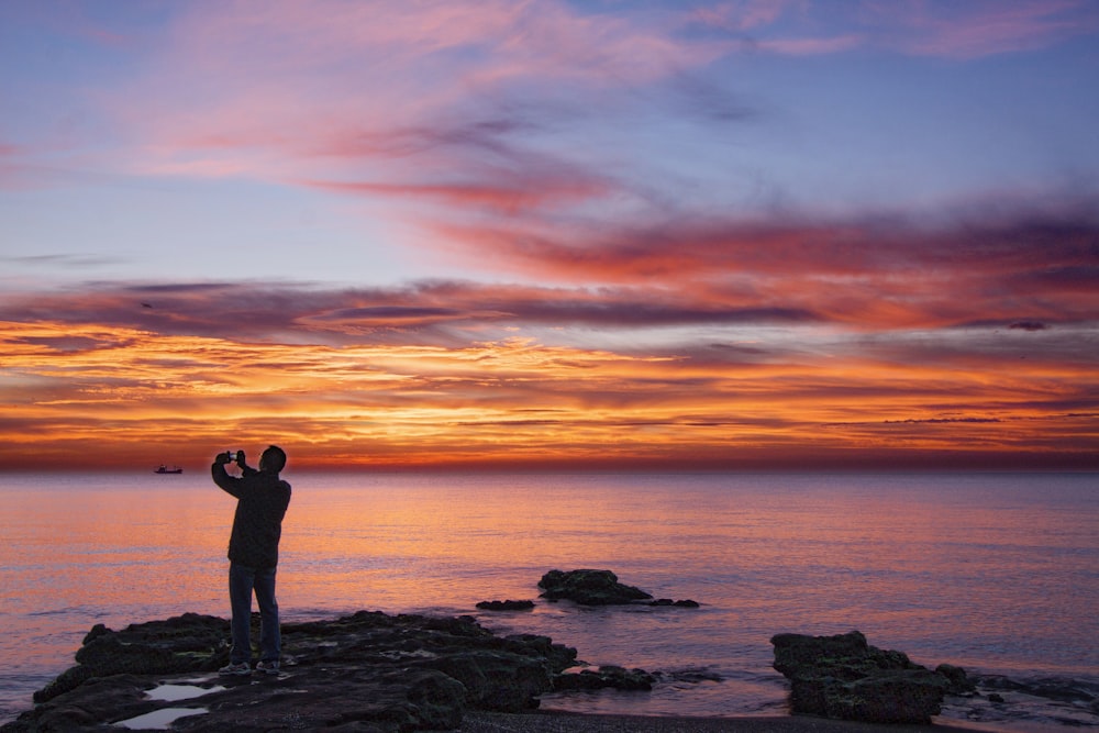 silhouette of woman standing on rock formation near body of water during sunset