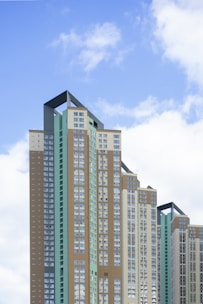 A group of modern high-rise buildings with a geometric design featuring various shades of brown, green, and beige against a blue sky. The buildings have numerous windows, and their facades include angular architectural elements at their tops.
